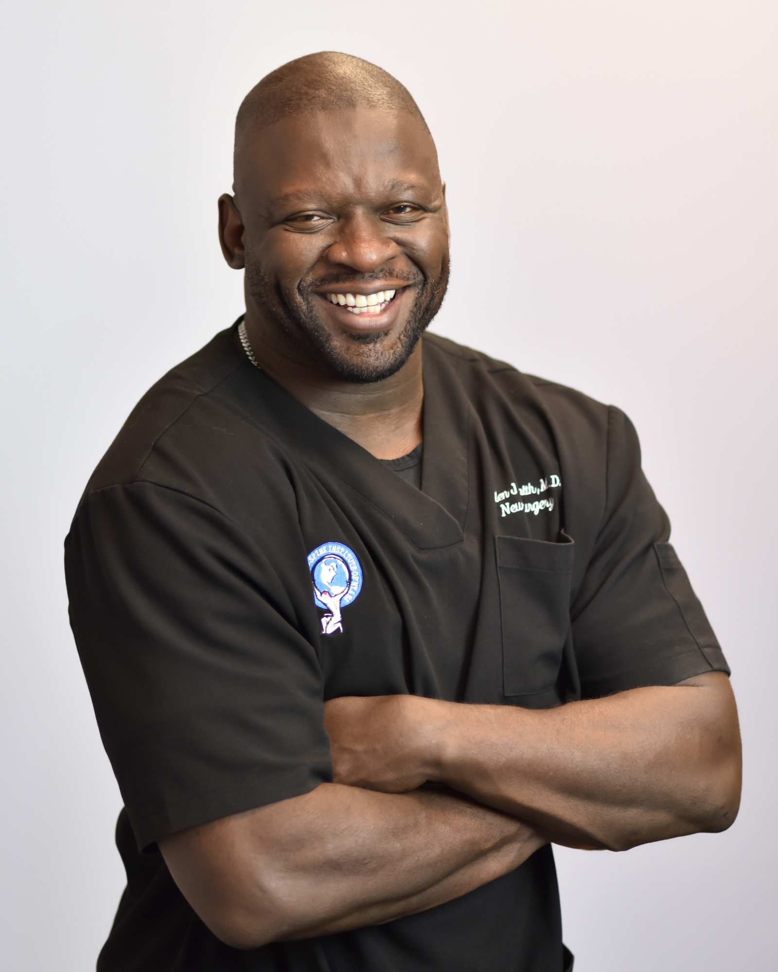 The image shows a man in a professional medical setting, wearing scrubs and smiling at the camera.