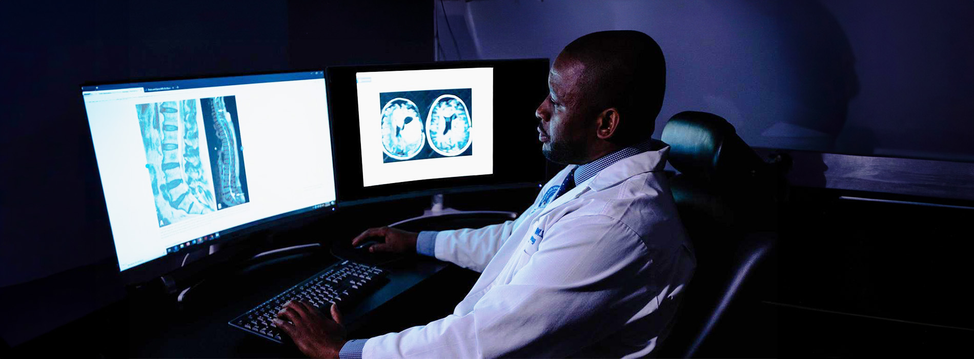A man sits at a computer with medical imaging displayed on multiple screens behind him.
