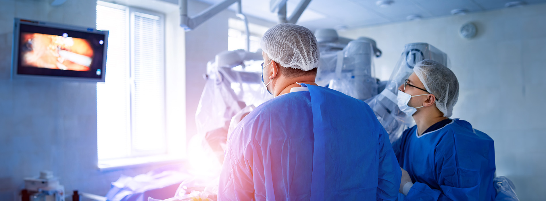 Two medical professionals in surgical attire standing in an operating room with medical equipment and screens in the background.